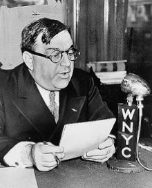 A man with glasses, holding a paper, speaking into a microphone